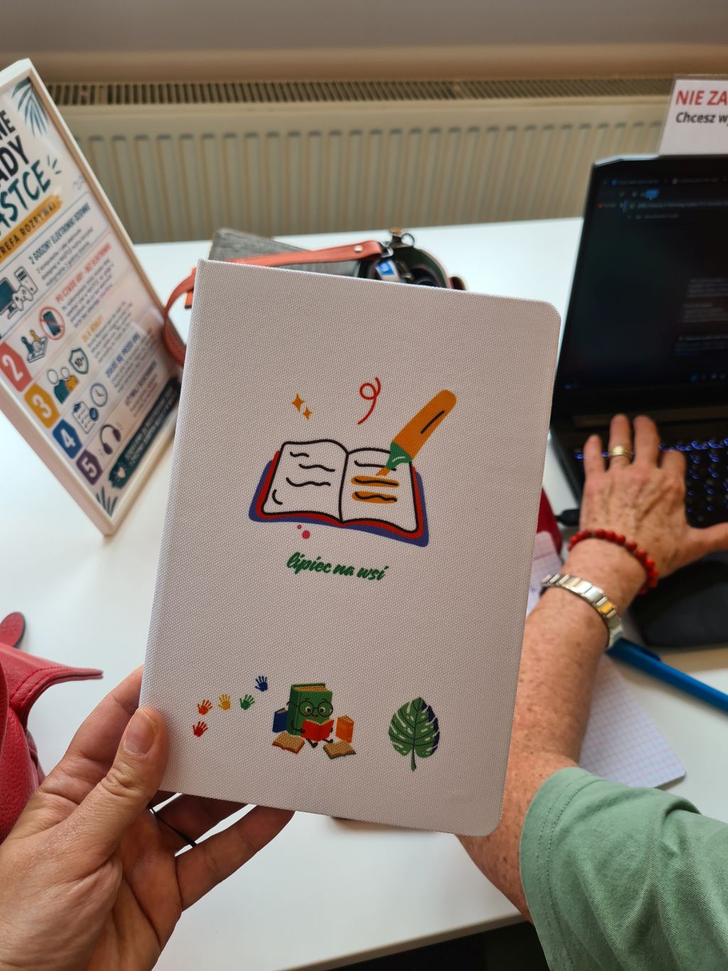 Uczestniczka prezentuje notes ozdobiony własnym projektem z grafikami książek, roślin i napisem „Otwórz na coś!”. W tle druga osoba pracuje przy laptopie.