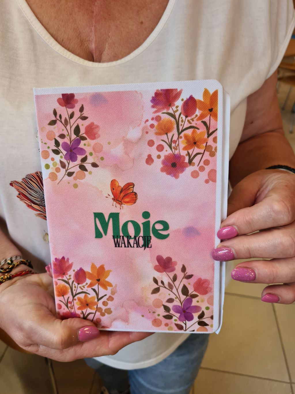 Uczestniczka trzyma notes z różową, kwiatową okładką, motylem i napisem „Moje wakacje”.