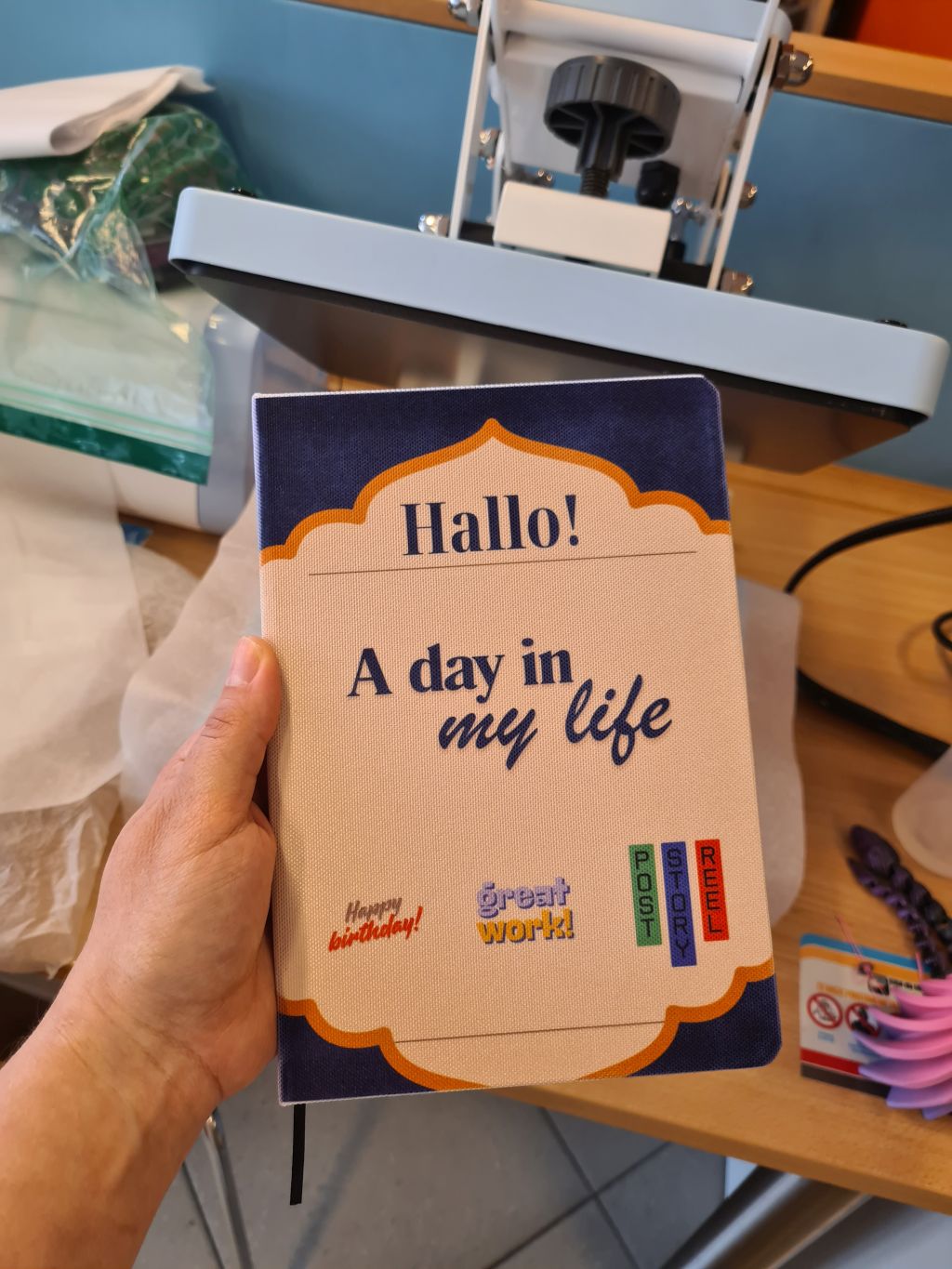 Dłoń trzyma notes z granatowo-beżową okładką i napisem „Hallo! A day in my life”. W tle widoczna jest prasa termiczna używana do nanoszenia projektów.