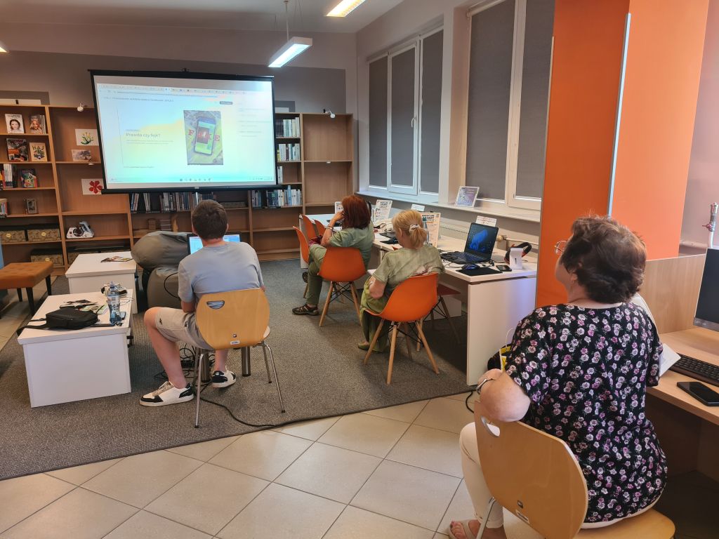 Uczestniczki warsztatów „Technosenior” siedzą przy stanowiskach komputerowych i oglądają prezentację wyświetlaną na dużym ekranie. Zajęcia prowadzi młodzieżowy lider Szymon