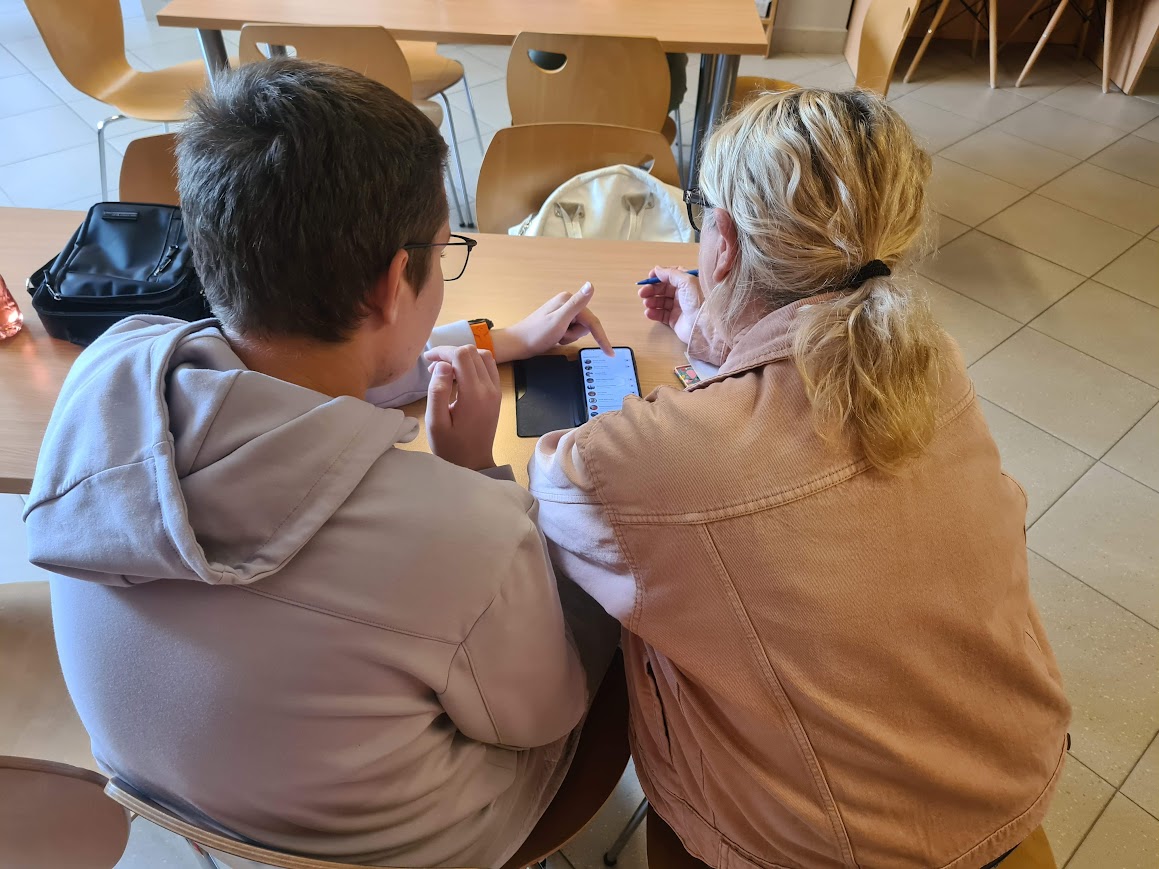 Młody mężczyzna siedzi przy stole obok seniorki i pokazuje jej funkcje telefonu. Oboje patrzą na ekran smartfona podczas indywidualnych konsultacji technologicznych.