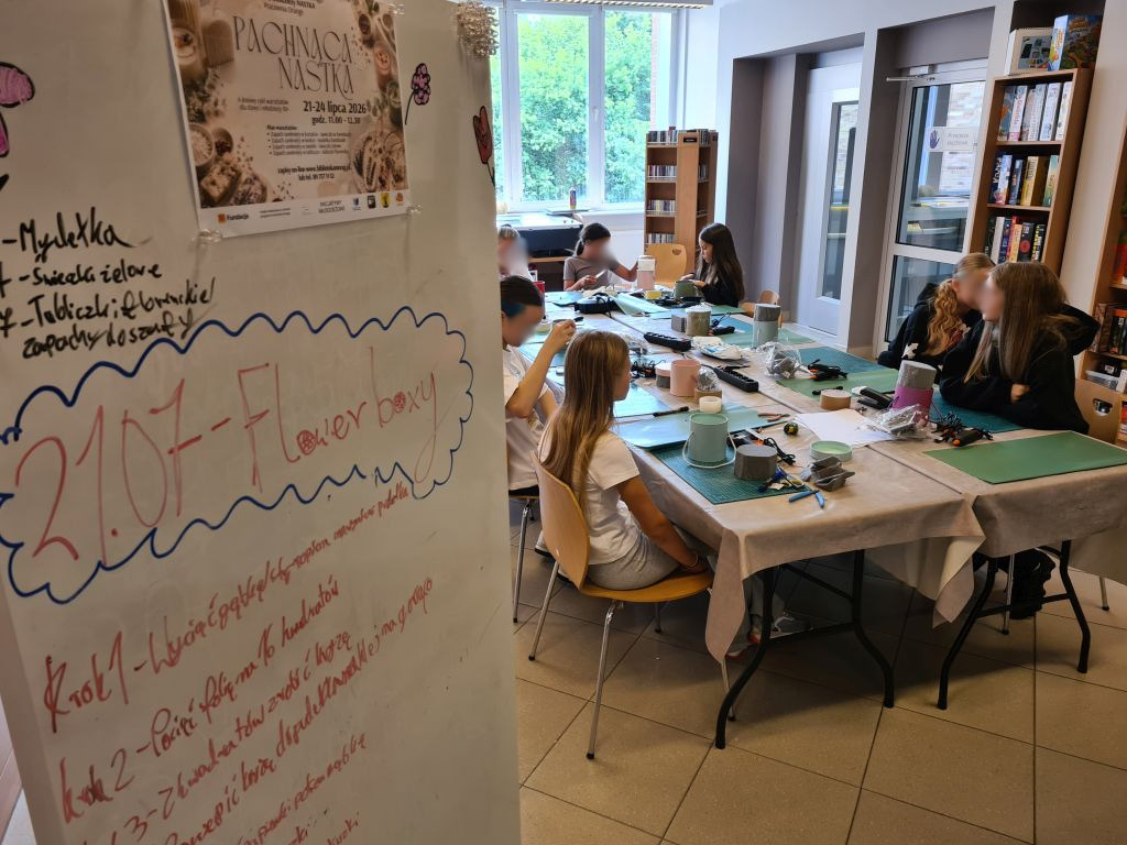 Uczestniczki warsztatów „Pachnąca NASTKA” pracują przy stole. Na pierwszym planie znajduje się tablica z planem zajęć, a w tle widoczny jest plakat wydarzenia.