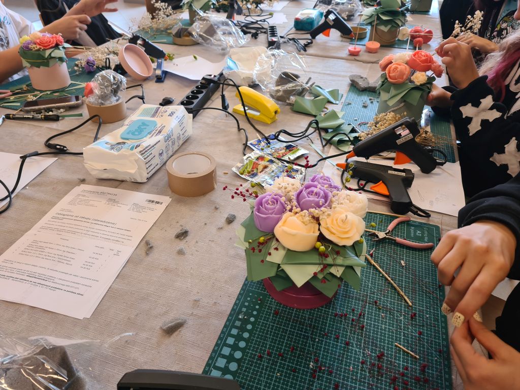 Kolorowy flowerbox ze świecami w kształcie róż i piwonii podczas wykonywania. Na stole leżą pistolety do kleju, nożyczki, suszone rośliny i pozostałe materiały florystyczne.