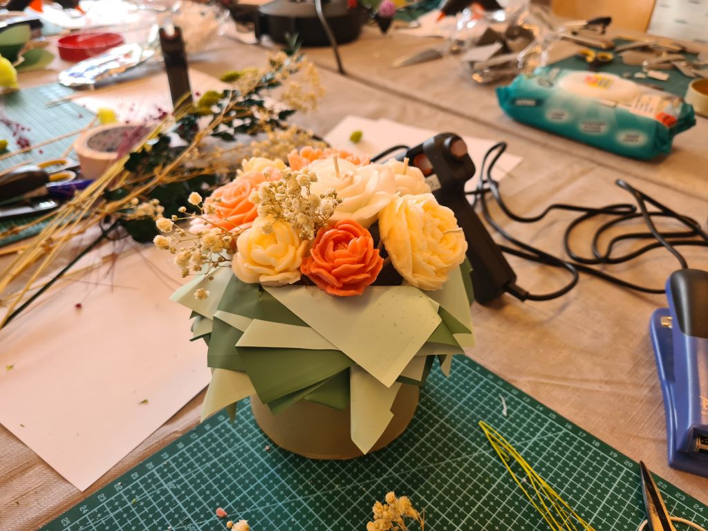 Gotowy flowerbox z kremowymi, pomarańczowymi i żółtymi świecami w kształcie kwiatów, ozdobiony gipsówką i zielonymi liśćmi.
