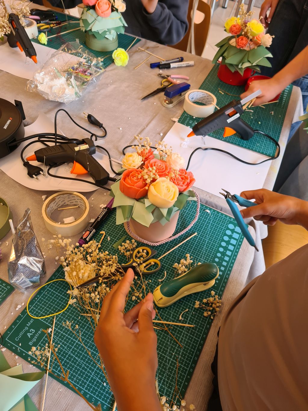 Uczestniczki dekorują pastelowy flowerbox świecami w kształcie róż. Na stole leżą pistolety do kleju, nożyczki, taśma i suszona gipsówka.