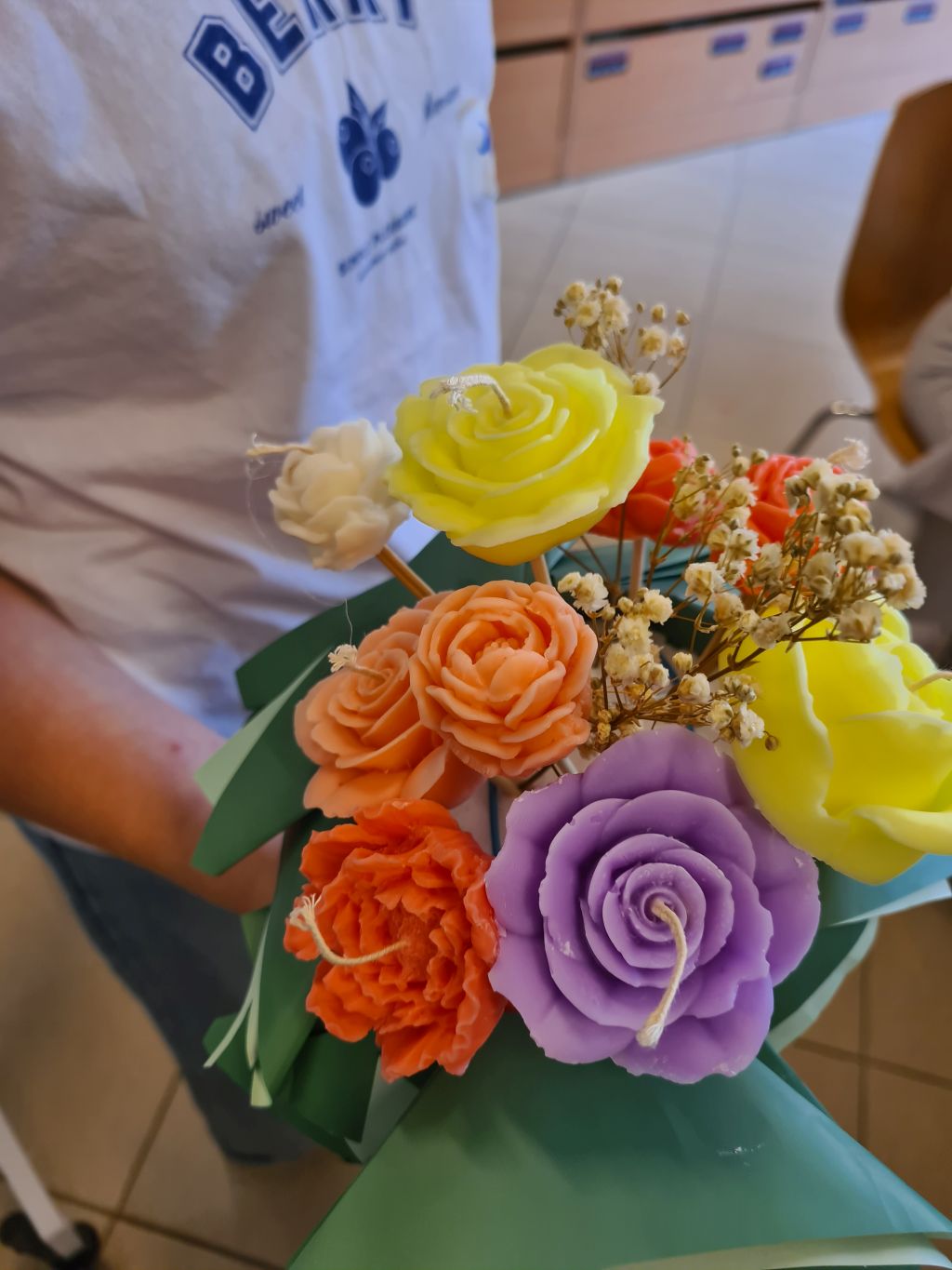 Gotowy flowerbox z kremowymi, pomarańczowymi i żółtymi świecami w kształcie kwiatów, ozdobiony gipsówką i zielonymi liśćmi.