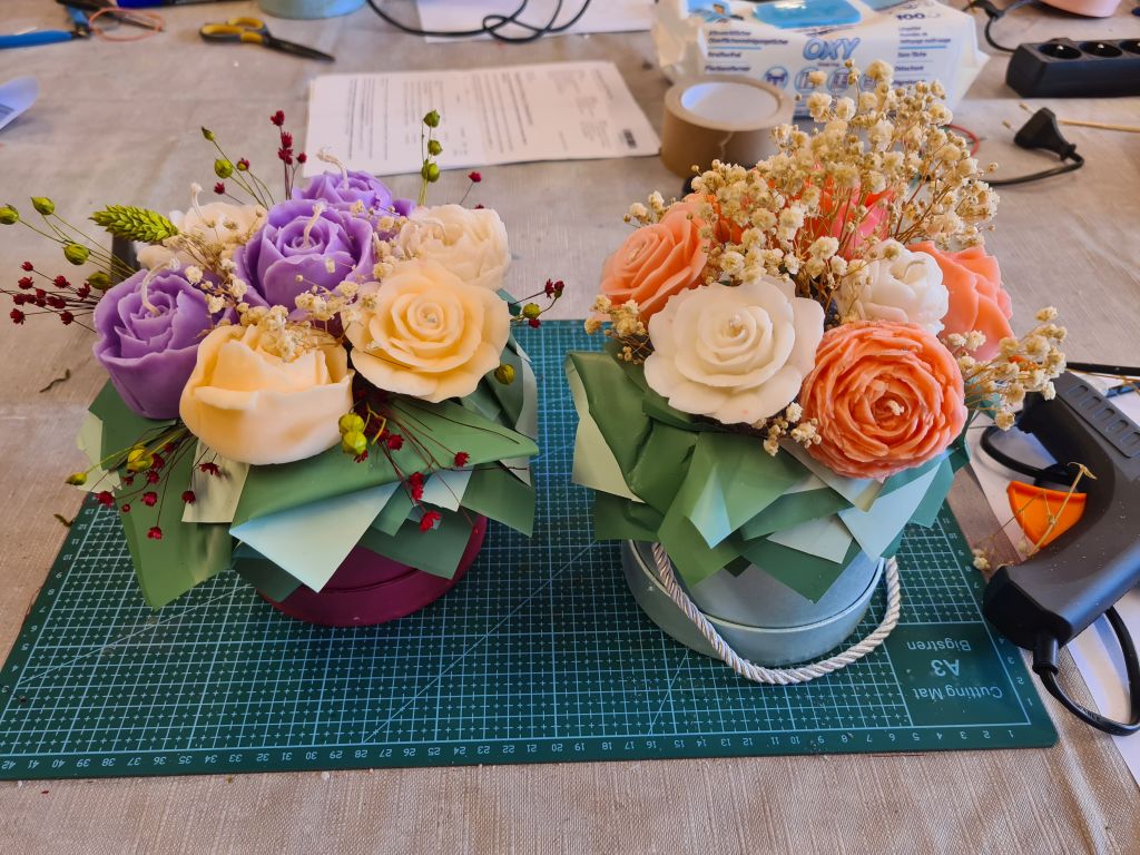 Dwa gotowe flowerboxy stoją obok siebie na macie do cięcia. Kompozycje zawierają fioletowe, kremowe, białe i koralowe świece oraz suszone kwiaty.