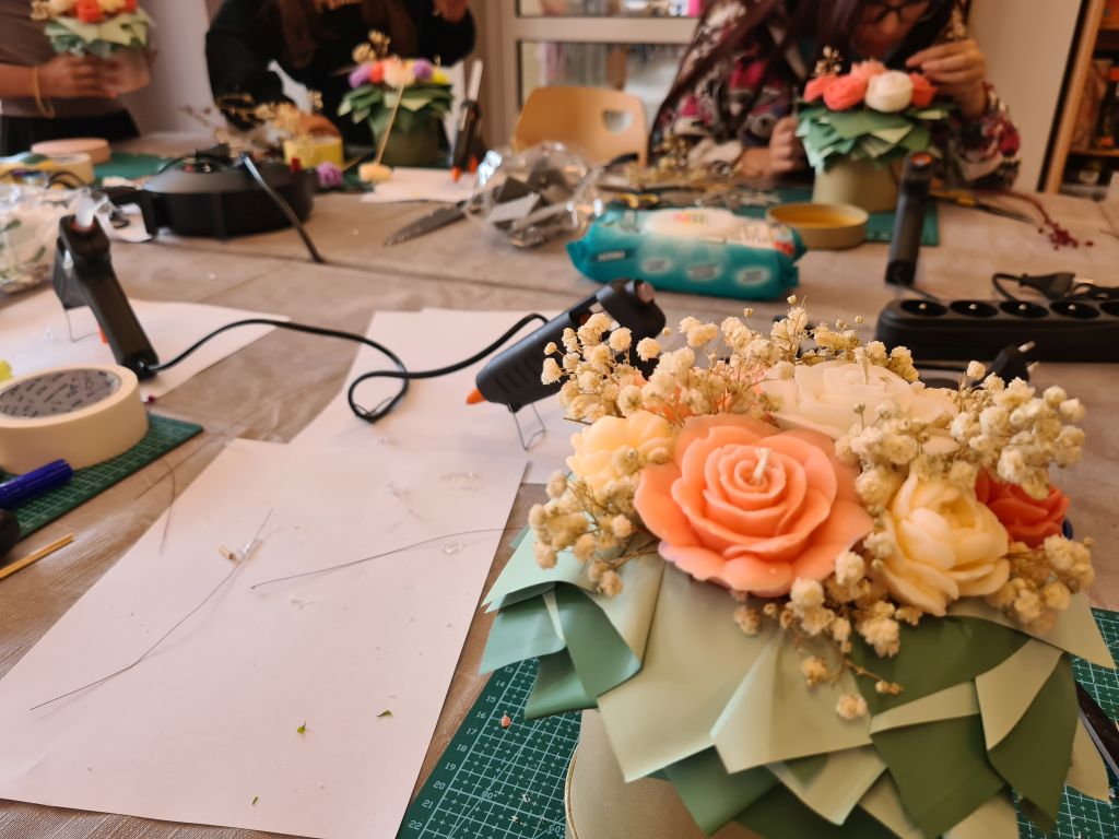 Gotowy flowerbox z koralowymi i kremowymi świecami w kształcie róż, bogato ozdobiony jasną gipsówką i zielonymi papierowymi liśćmi.