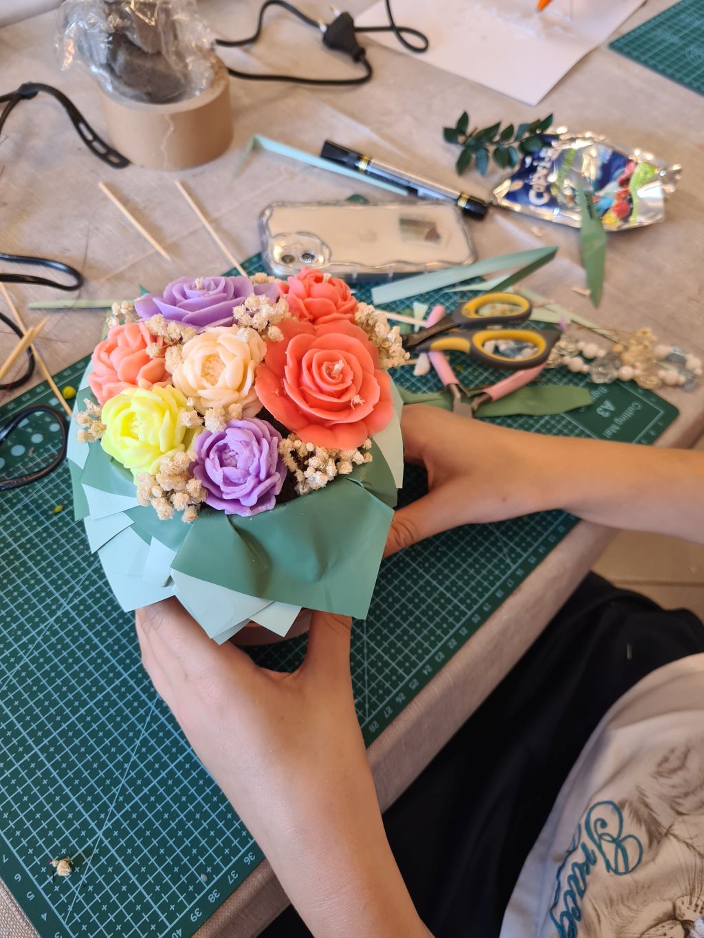 Uczestniczka trzyma pastelowy flowerbox z różowymi, fioletowymi, kremowymi i żółtymi świecami-kwiatami. W tle leżą nożyczki, szczypce i materiały dekoracyjne.