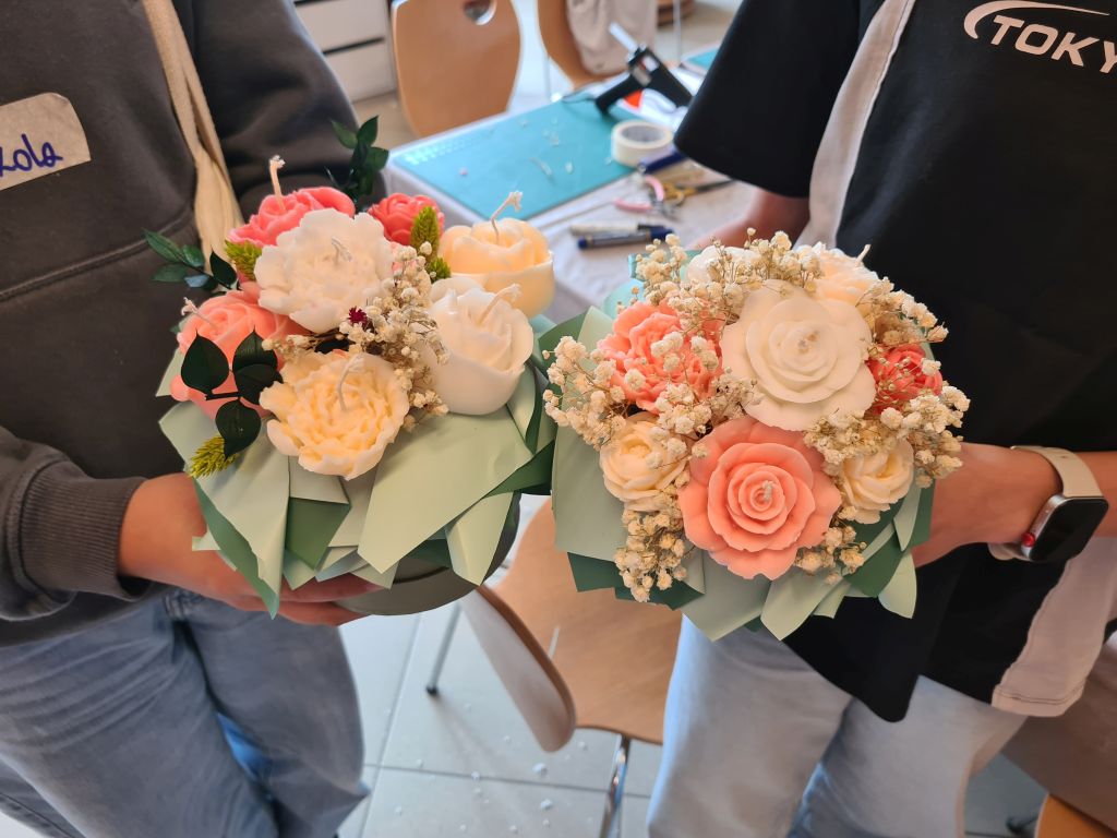 Dwie uczestniczki prezentują wykonane flowerboxy z białymi, kremowymi i koralowymi świecami oraz jasną gipsówką.