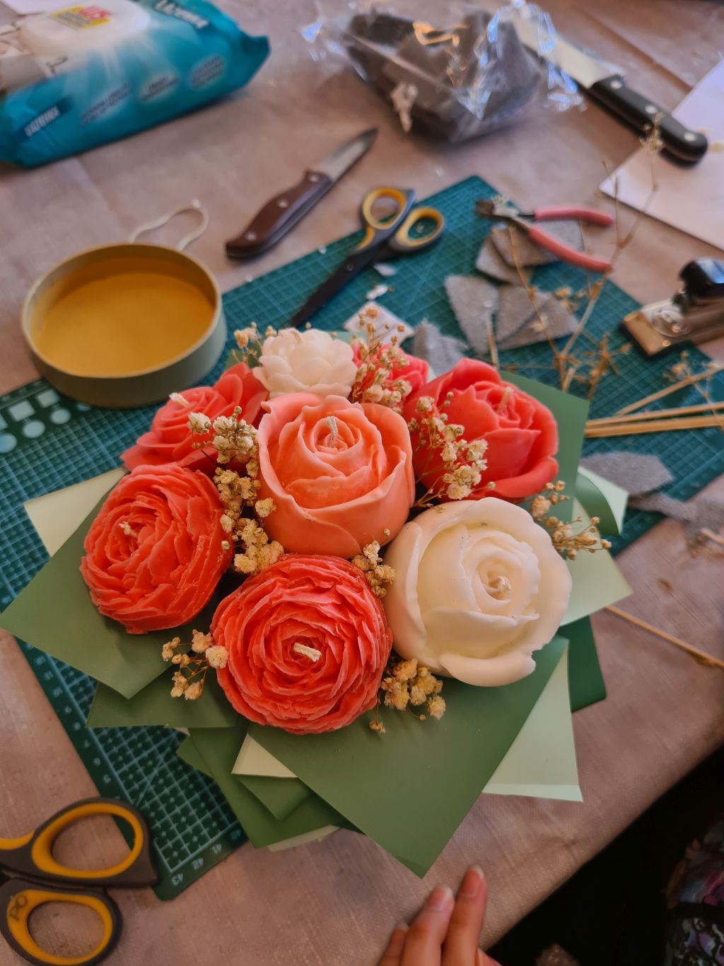 Gotowy flowerbox z koralowymi i kremowymi świecami w kształcie róż i piwonii. Kompozycję uzupełniają drobne jasne kwiaty oraz papierowe liście w kilku odcieniach zieleni.