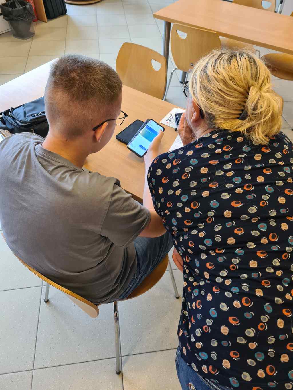 Młody mężczyzna pomaga seniorce w obsłudze smartfona. Uczestnicy siedzą obok siebie przy stole i wspólnie analizują informacje widoczne na ekranie telefonu.