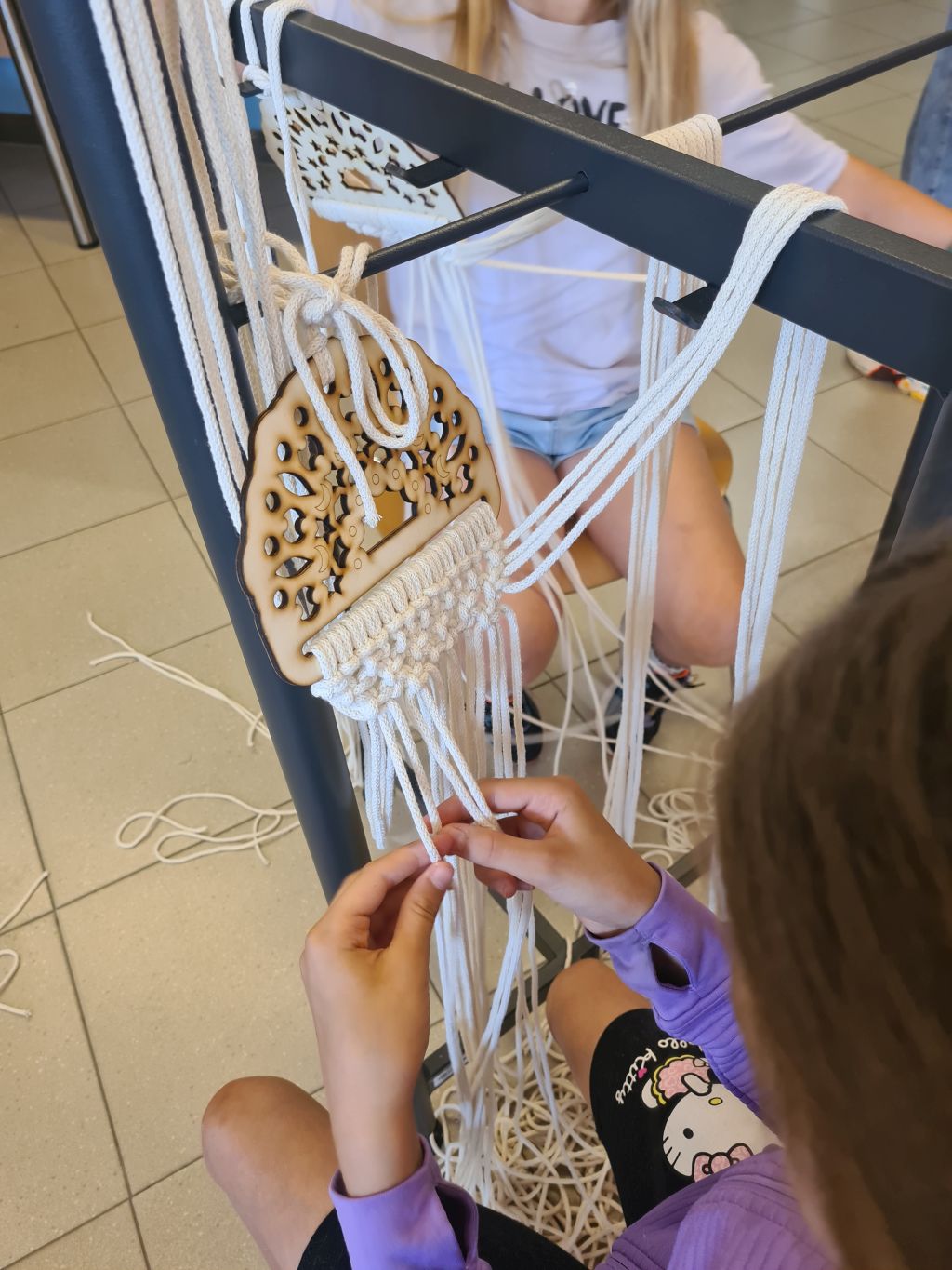 Zbliżenie na dłonie uczestniczki wykonującej kolejne węzły makramy pod ozdobną, drewnianą bazą.