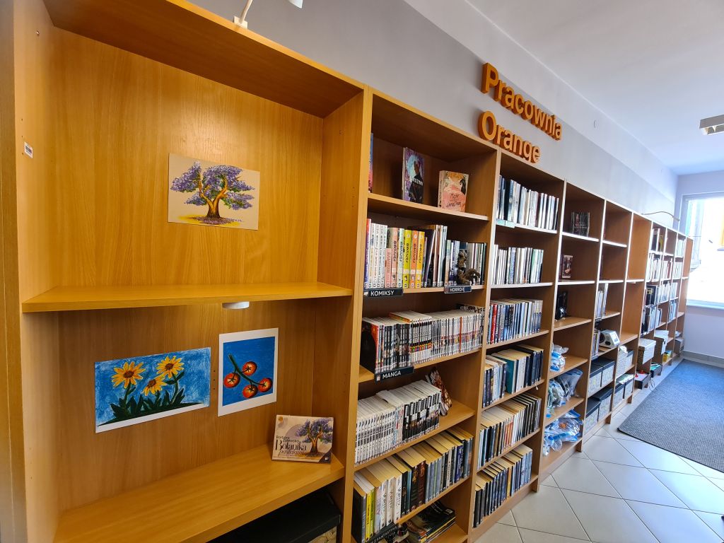 Fragment Galerii Między Książkami w Dziale dla Młodzieży NASTKA. Na drewnianych regałach, obok książek i napisu „Pracownia Orange”, wyeksponowano pastelowe prace przedstawiające drzewo, słoneczniki i owoce.