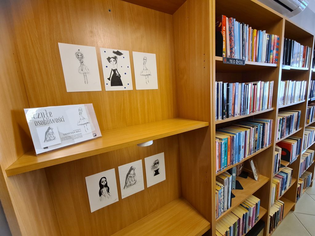 Fragment wystawy „Czułe osobliwości” wśród bibliotecznych regałów. Na drewnianej ściance rozmieszczono sześć czarno-białych rysunków smukłych, fantastycznych postaci, a na półce stoi tabliczka z tytułem wystawy i nazwiskiem artystki.