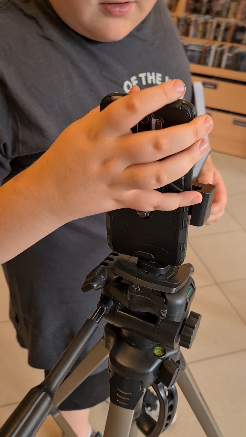 Zbliżenie na dłonie osoby w ciemnej koszulce, która montuje lub dopasowuje smartfon w czarnym, masywnym etui na statywie fotograficznym.