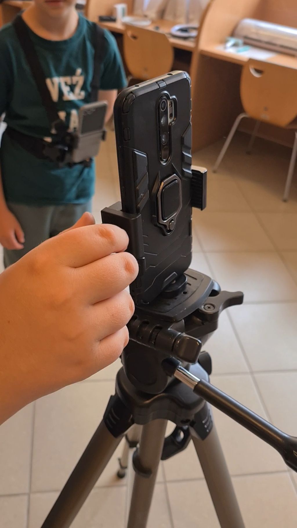 Dłoń regulująca uchwyt smartfona zamocowanego pionowo na statywie. W rozmytym tle widać stojącą postać w zielonej koszulce oraz wnętrze pokoju z biurkami.
