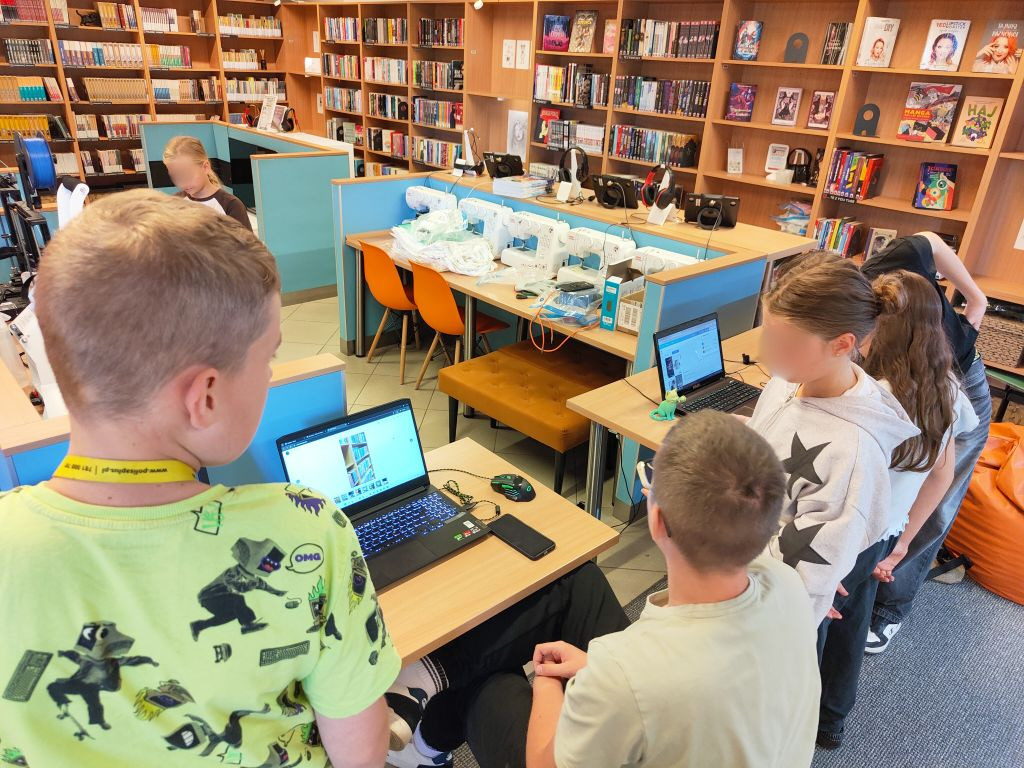 Dzieci i młodzież skupieni wokół stanowiska komputerowego patrzą na ekran laptopa. W tle widać regały z książkami oraz maszyny do szycia.