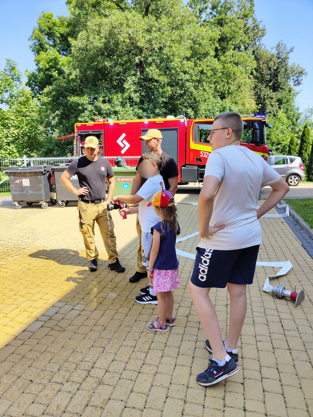 Strażak prezentuje dzieciom prądnicę pożarniczą podłączoną do węża na wybrukowanym placu przed czerwonym wozem strażackim.