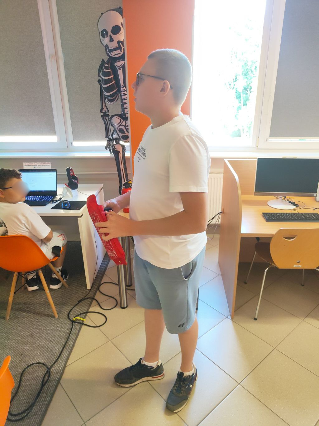 Chłopiec w okularach i białej koszulce trzymający w rękach czerwoną gaśnicę w sali komputerowej.