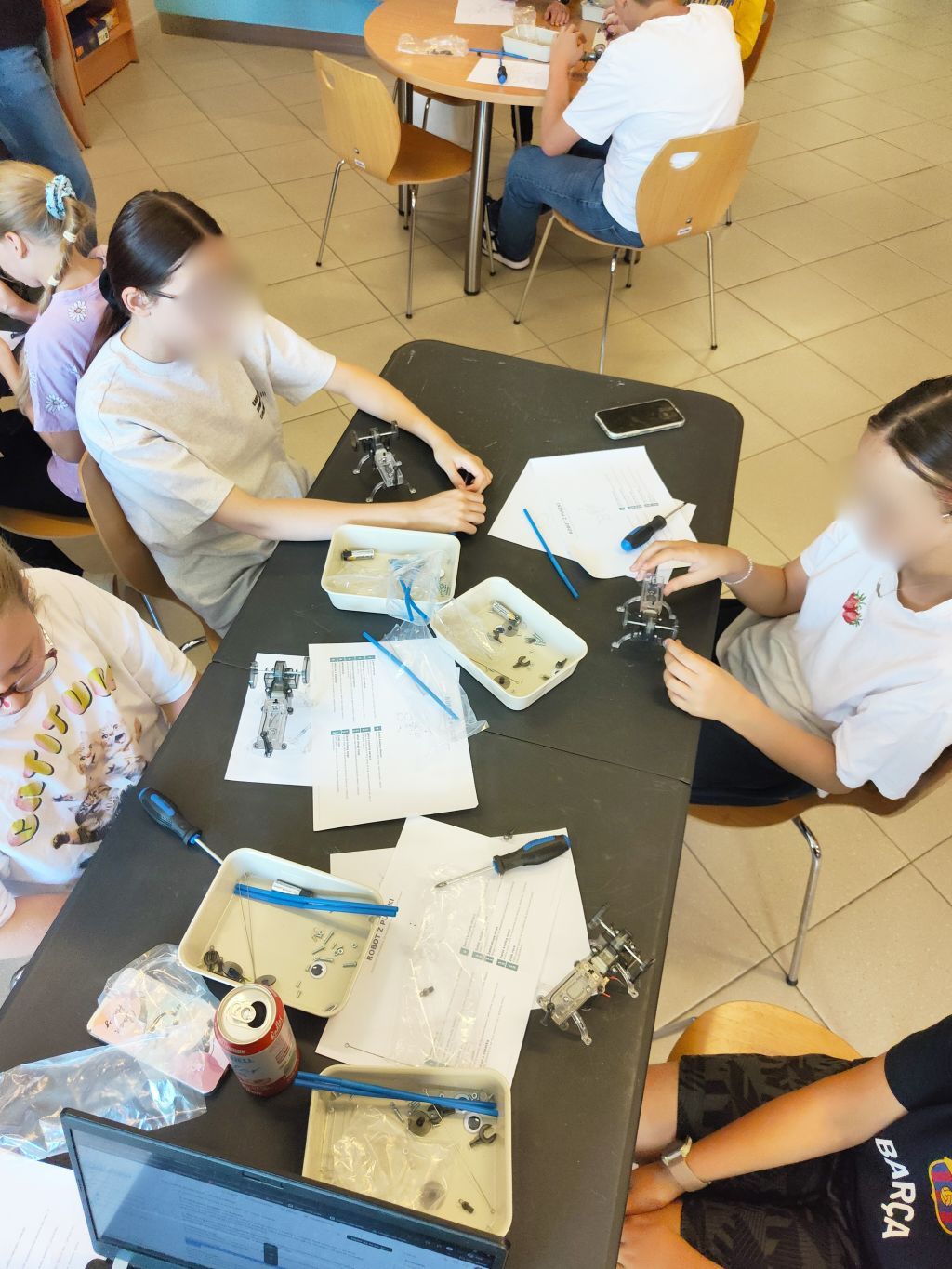Widok z góry na trzy dziewczyny siedzące przy czarnym stole, składające zestawy robotów, z tacami, śrubokrętami, puszką i instrukcjami rozłożonymi przed nimi.