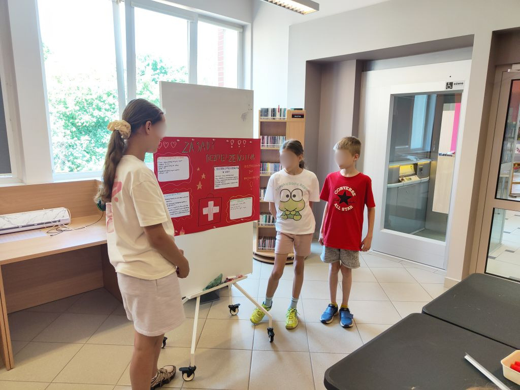 Troje dzieci stojących przy białej tablicy z czerwoną planszą o nazwie „Zasady bezpieczeństwa”.
