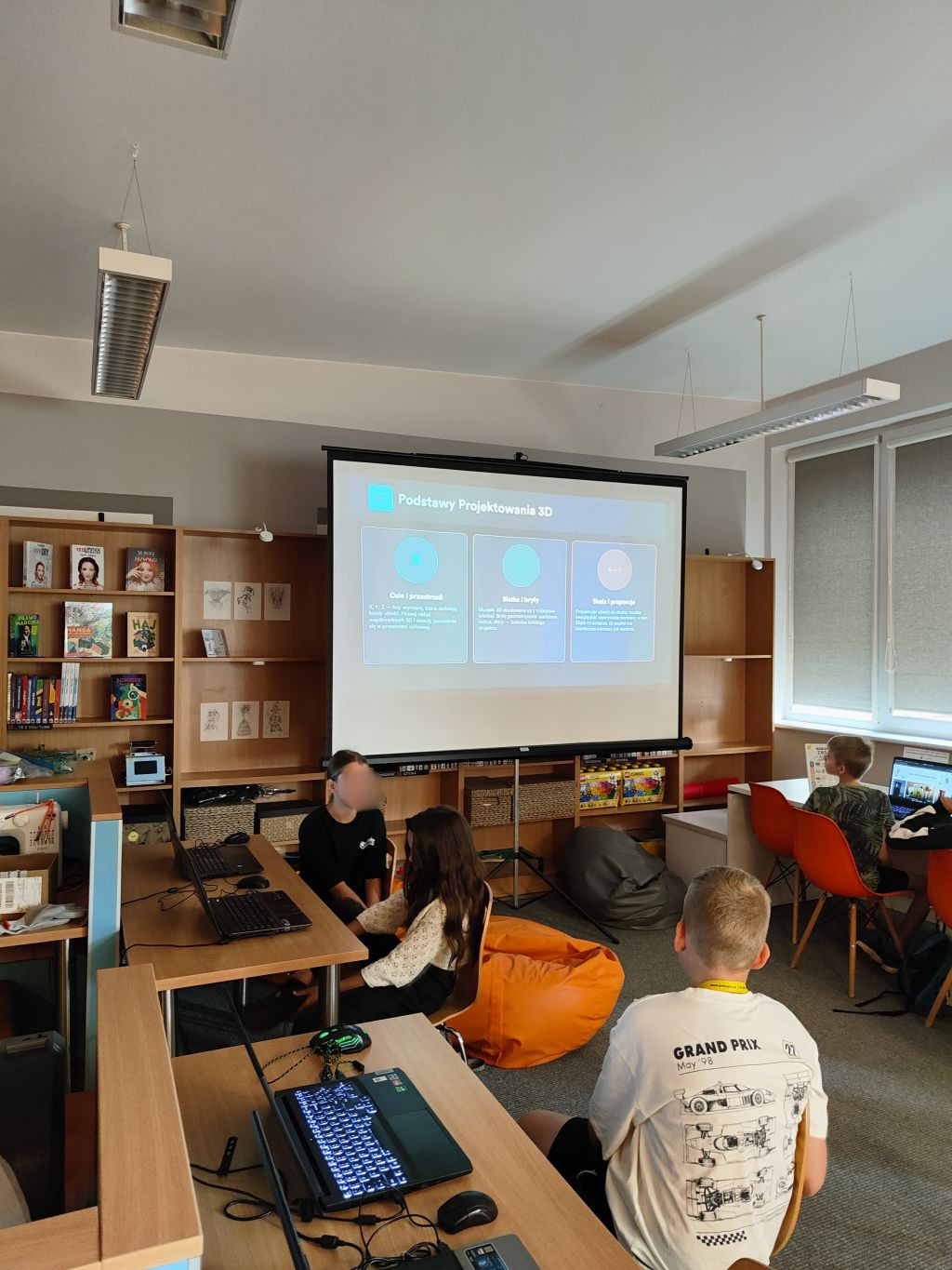 Uczestnicy zajęć słuchają prezentacji pt. „Podstawy Projektowania 3D” wyświetlanej na dużym ekranie projekcyjnym w sali z regałami pełnymi książek.