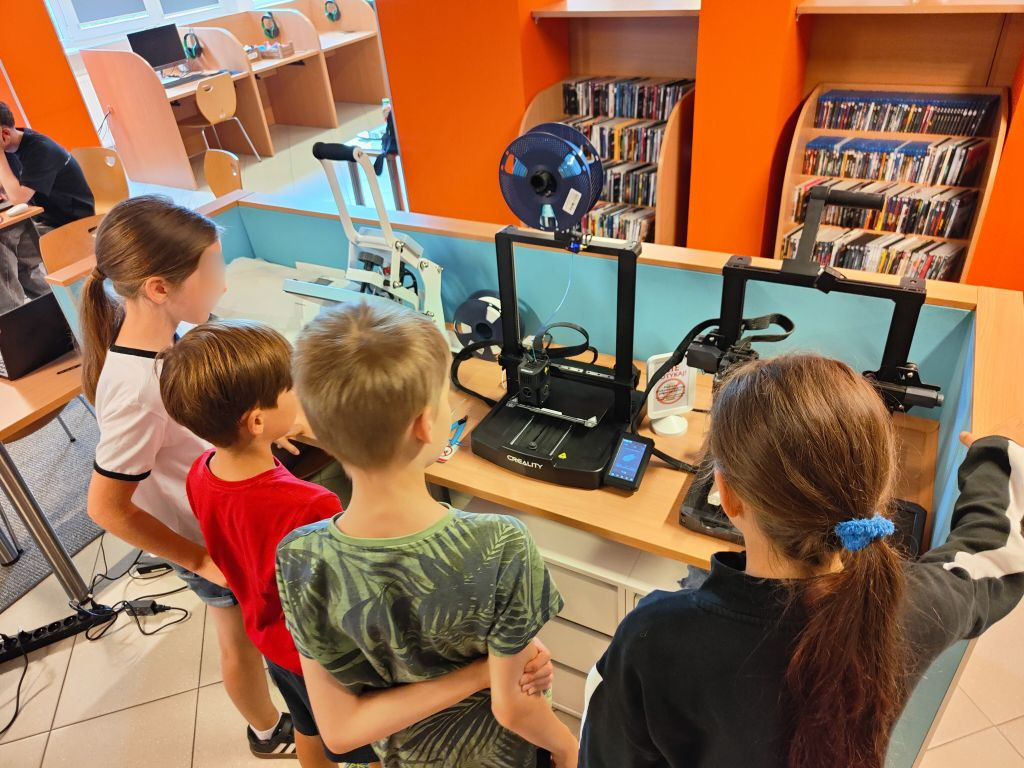 Czworo dzieci stoi obok stoiska z pracującymi drukarkami 3D i przygląda się procesowi druku.