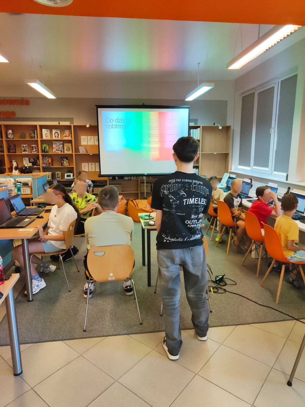 Chłopak w czarnej koszulce stoi tyłem przed ekranem projekcyjnym z napisem „Co dzisiaj robimy?”. Wokół niego dzieci siedzą przy stołach i pracują na laptopach.