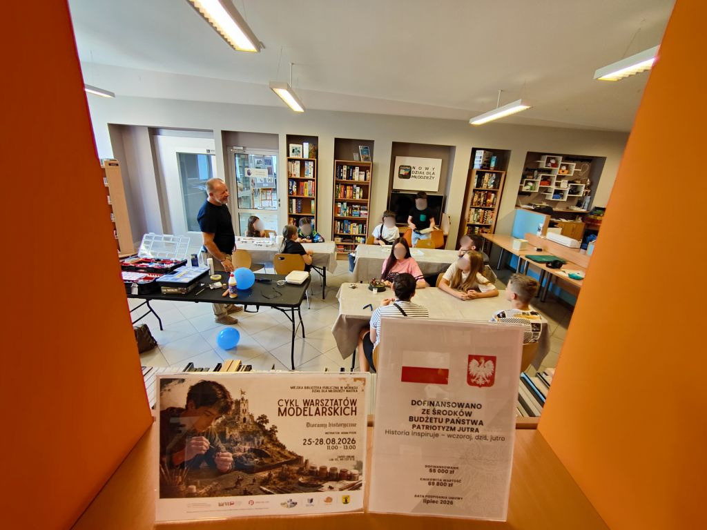 Ogólny widok sali bibliotecznej, w której grupa dzieci siedzi przy stołach i zajmuje się modelarstwem pod okiem instruktora. Na pierwszym planie widoczne są dwa plakaty informacyjne o warsztatach.