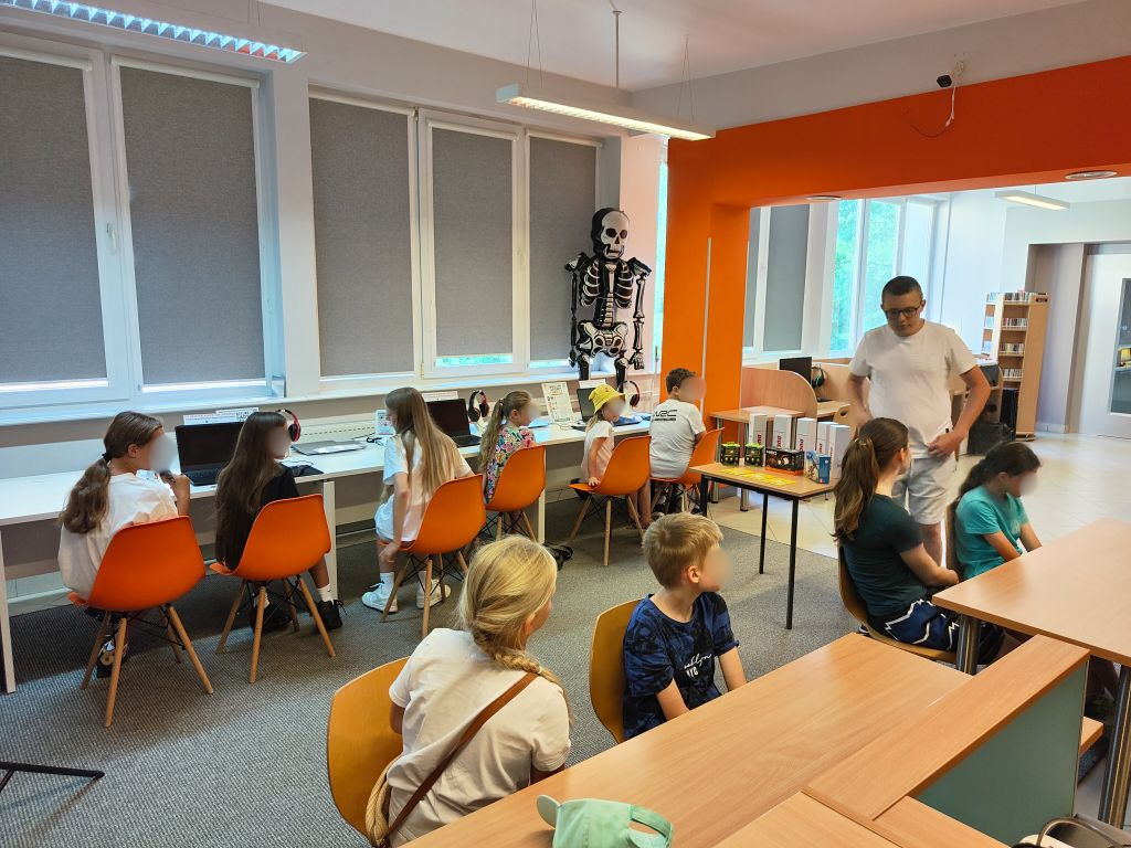 Prowadzący rozmawia z dziećmi siedzącymi przy biurkach i stolikach w jasnej sali bibliotecznej z widocznym w tle modelem szkieletu.