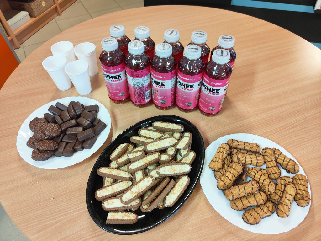 Przekąski na stole: ciastka na trzech talerzach, jednorazowe kubeczki oraz butelki napoju Oshee Vitamin Water.