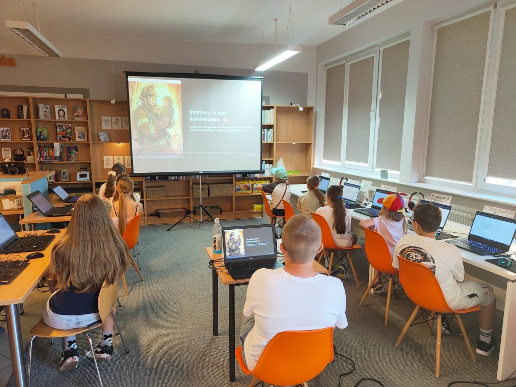 Dzieci siedzące przy biurkach z laptopami i oglądające prezentację o strażakach wyświetlaną na ekranie projekcyjnym.