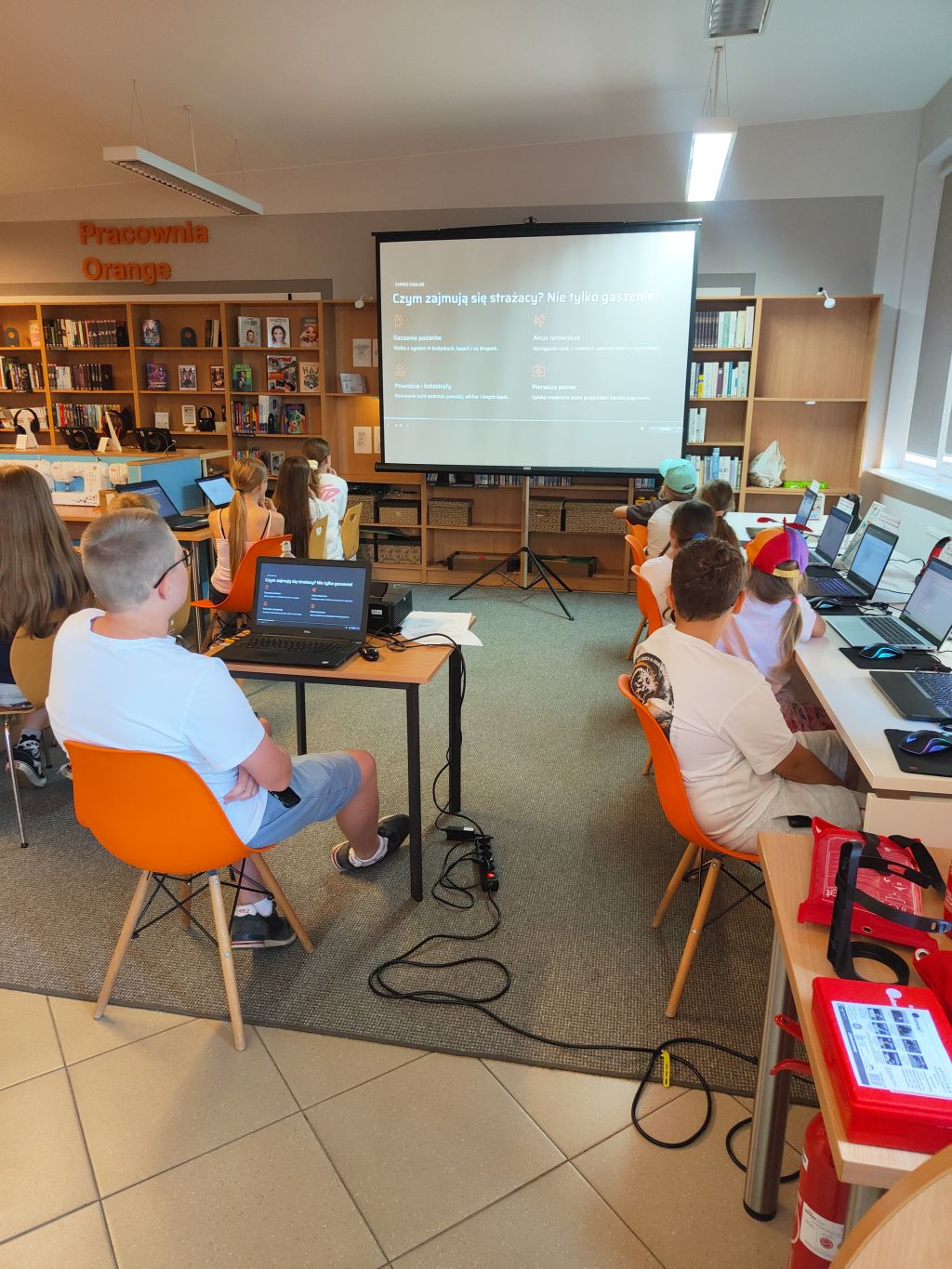 Dzieci siedzące przy stanowiskach z laptopami w bibliotece, oglądające prezentację na ekranie projekcyjnym.