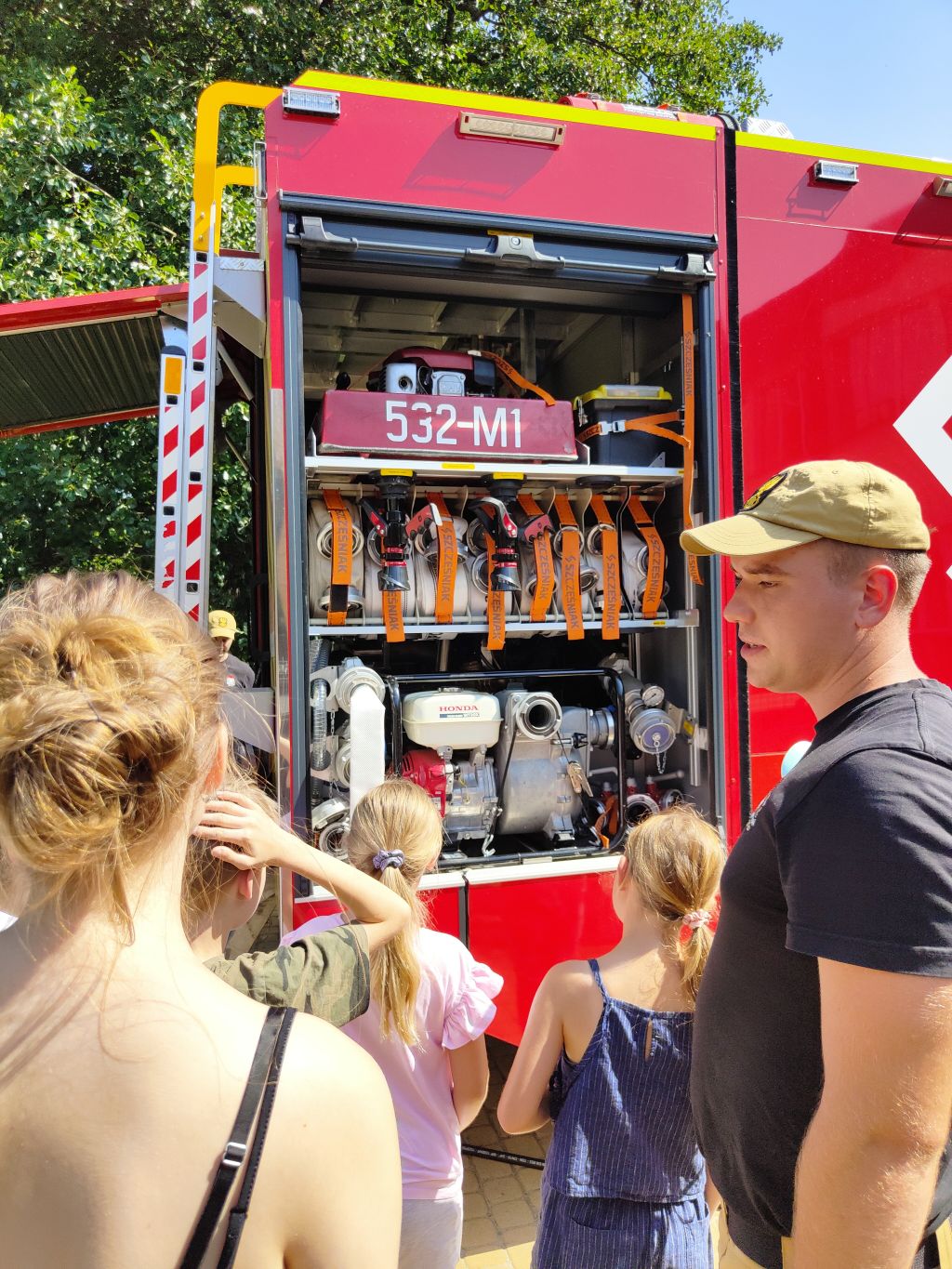 Strażak stoi z dziećmi przy otwartej skrytce wozu strażackiego, prezentując motopompę i zwinięte węże.