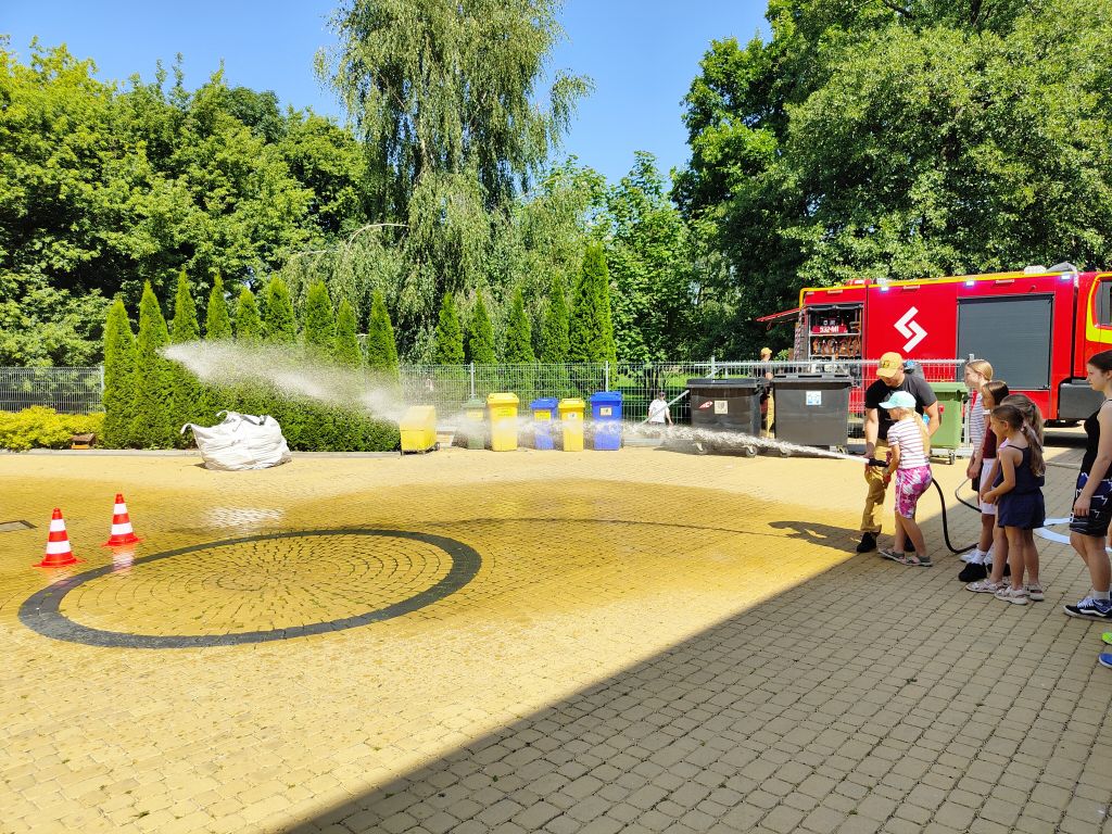Dziewczynka z pomocą strażaka celuje strumieniem wody z wężą w stronę pachołków na placu.