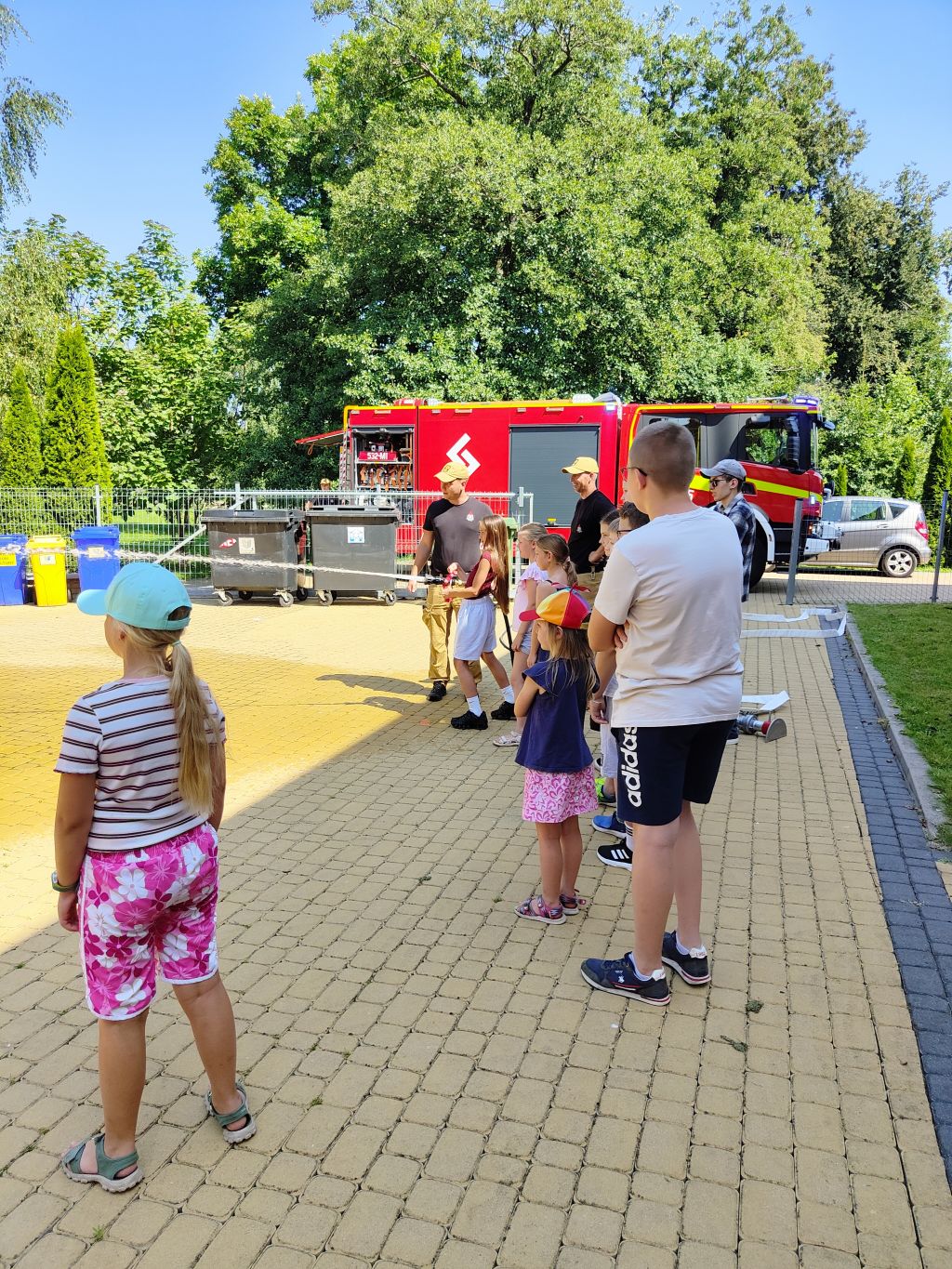 Grupa dzieci obserwuje pokaz lania wody z wężą strażackiego prowadzony przez strażaka na placu przed czerwonym wozem.
