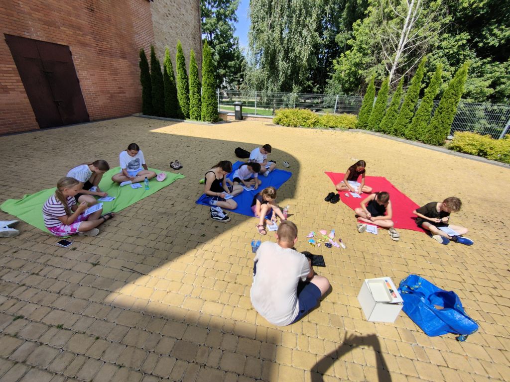 Dzieci i opiekun siedzą na kolorowych kocykach rozłożonych na kostce brukowej, wypełniając karty pracy na świeżym powietrzu.