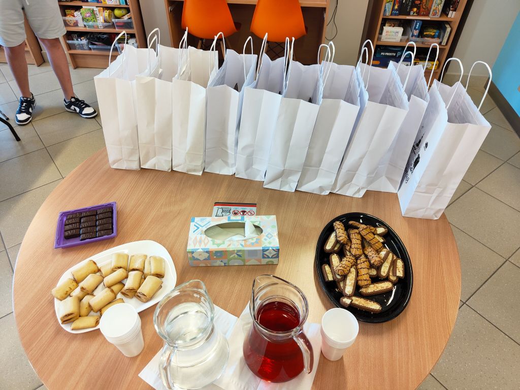 Na stoliku przygotowano poczęstunek w postaci ciastek i napojów oraz rząd białych toreb z upominkami dla uczestników.
