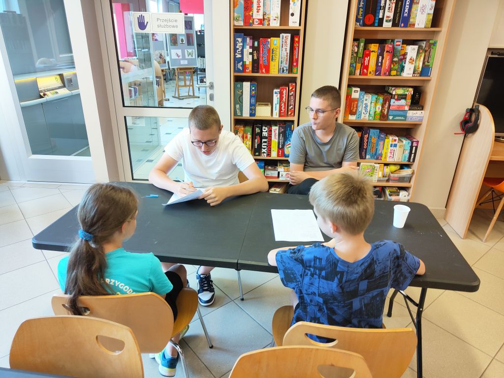 Dwójka dzieci siedzi przy stoliku naprzeciwko dwojga prowadzących, którzy uzupełniają arkusz odpowiedzi podczas quizu w bibliotece.