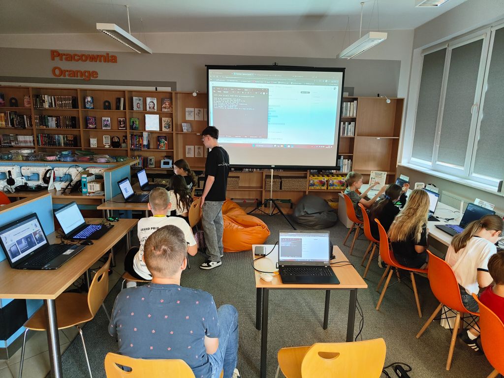 Dzieci pracują na laptopach przy stołach w sali komputerowej z regałami na książki, pod nadzorem instruktora stojącego przy ekranie projekcyjnym.