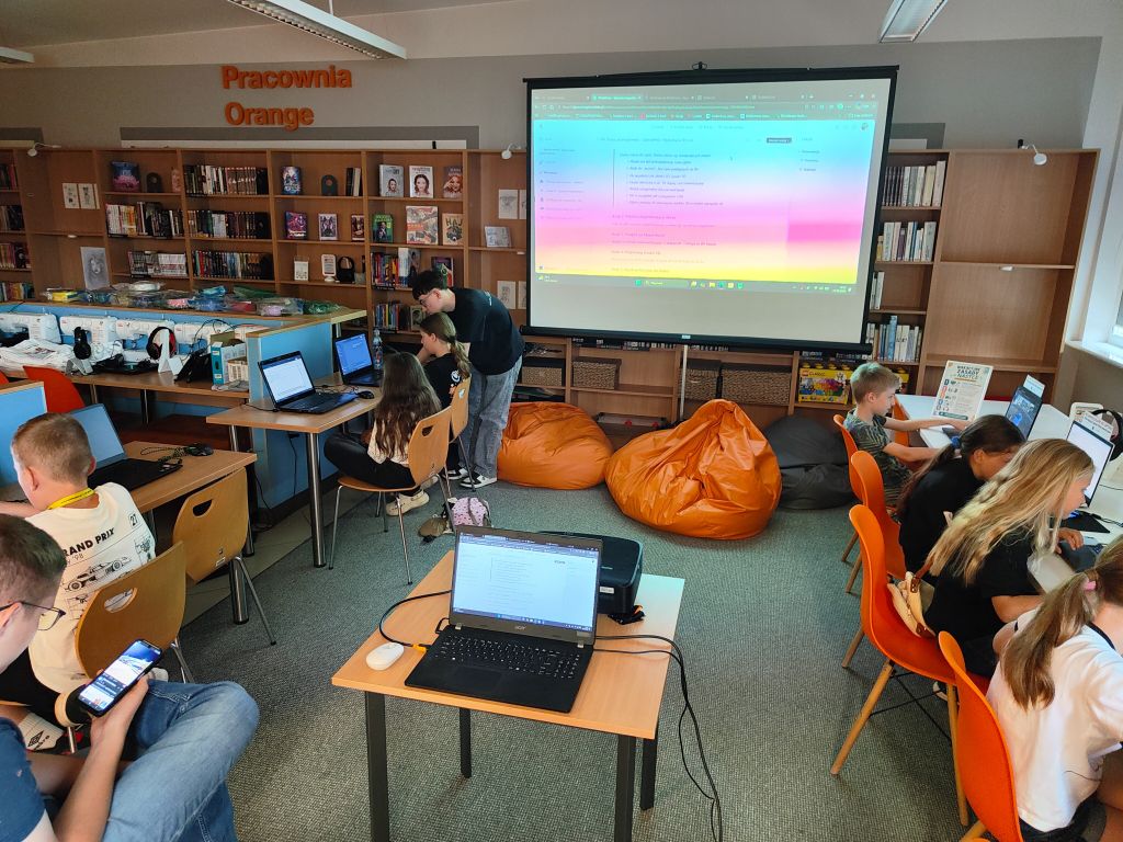 Szerokie ujęcie sali komputerowej „Pracownia Orange”, w której dzieci pracują na laptopach, a na ekranie wyświetlana jest prezentacja z kolorowym tłem.