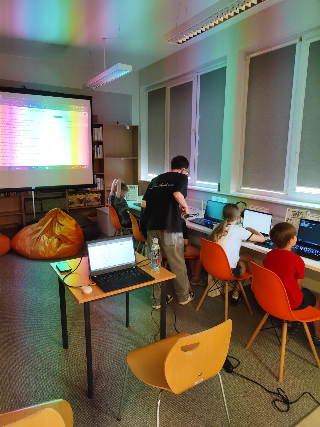 Instruktor pochyla się nad dzieckiem pracującym przy laptopie pod oknem w sali oświetlanej kolorowym światłem.