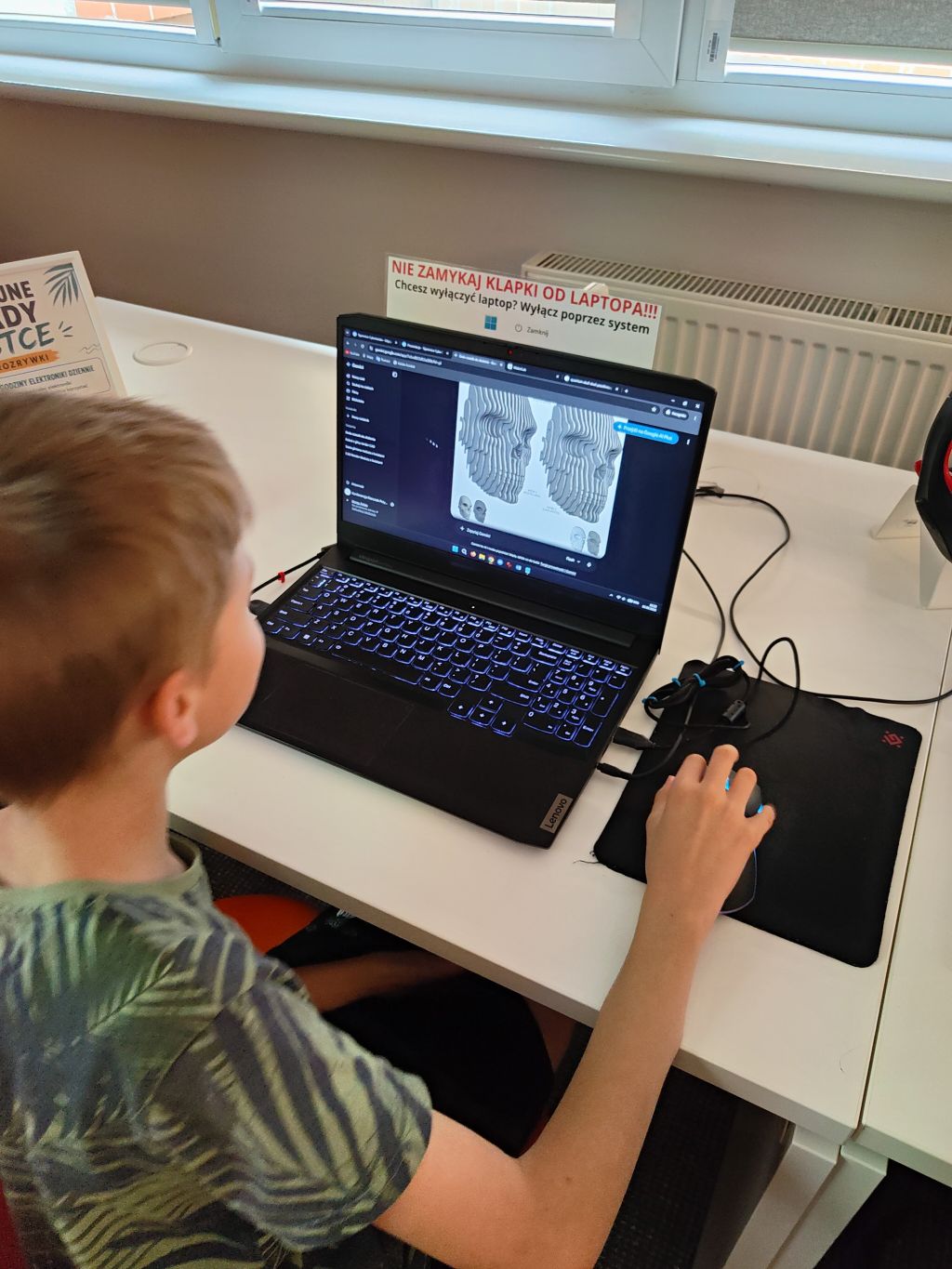Chłopiec siedzi przy białym biurku i obsługuje myszką komputer, na którego ekranie widać model rzeźby twarzy w programie 3D.