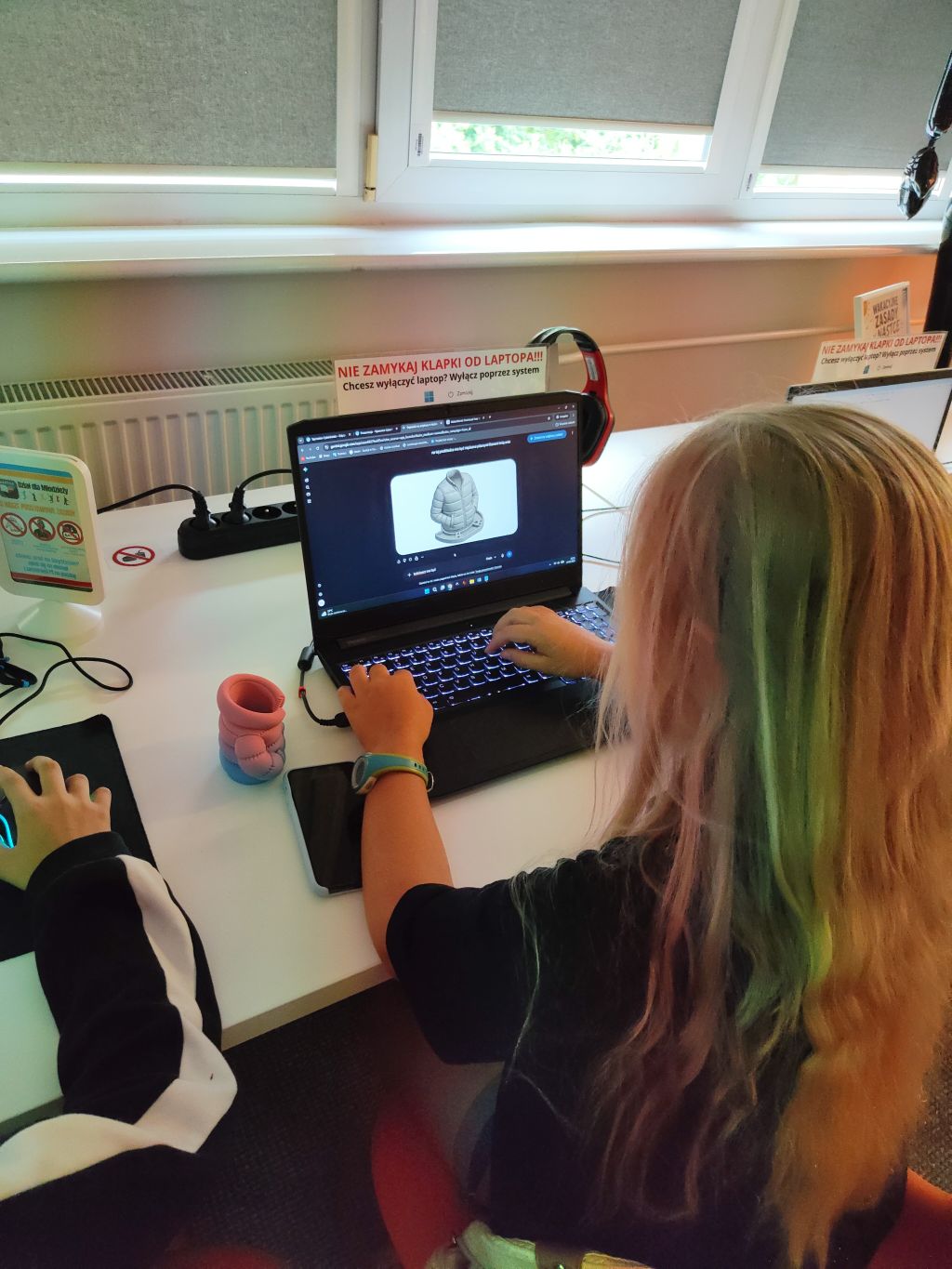 Widok zza pleców dziewczyny, która pracuje przy laptopie w nowoczesnej pracowni komputerowej, obok innej osoby używającej myszki.