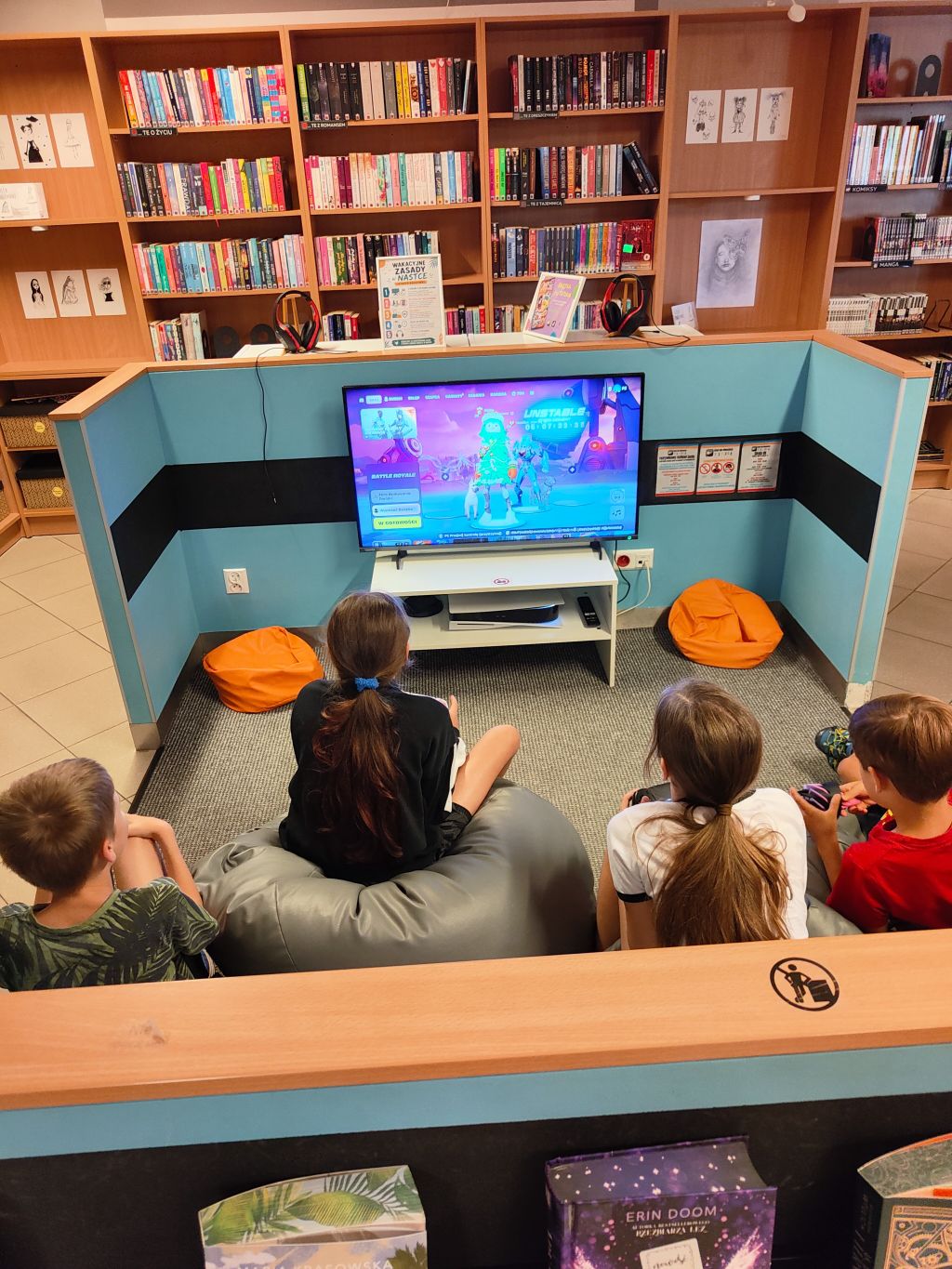 Czworo dzieci siedzi na pufach w specjalnej strefie z błękitnymi ściankami i gra w grę wideo wyświetlaną na dużym telewizorze w bibliotece.