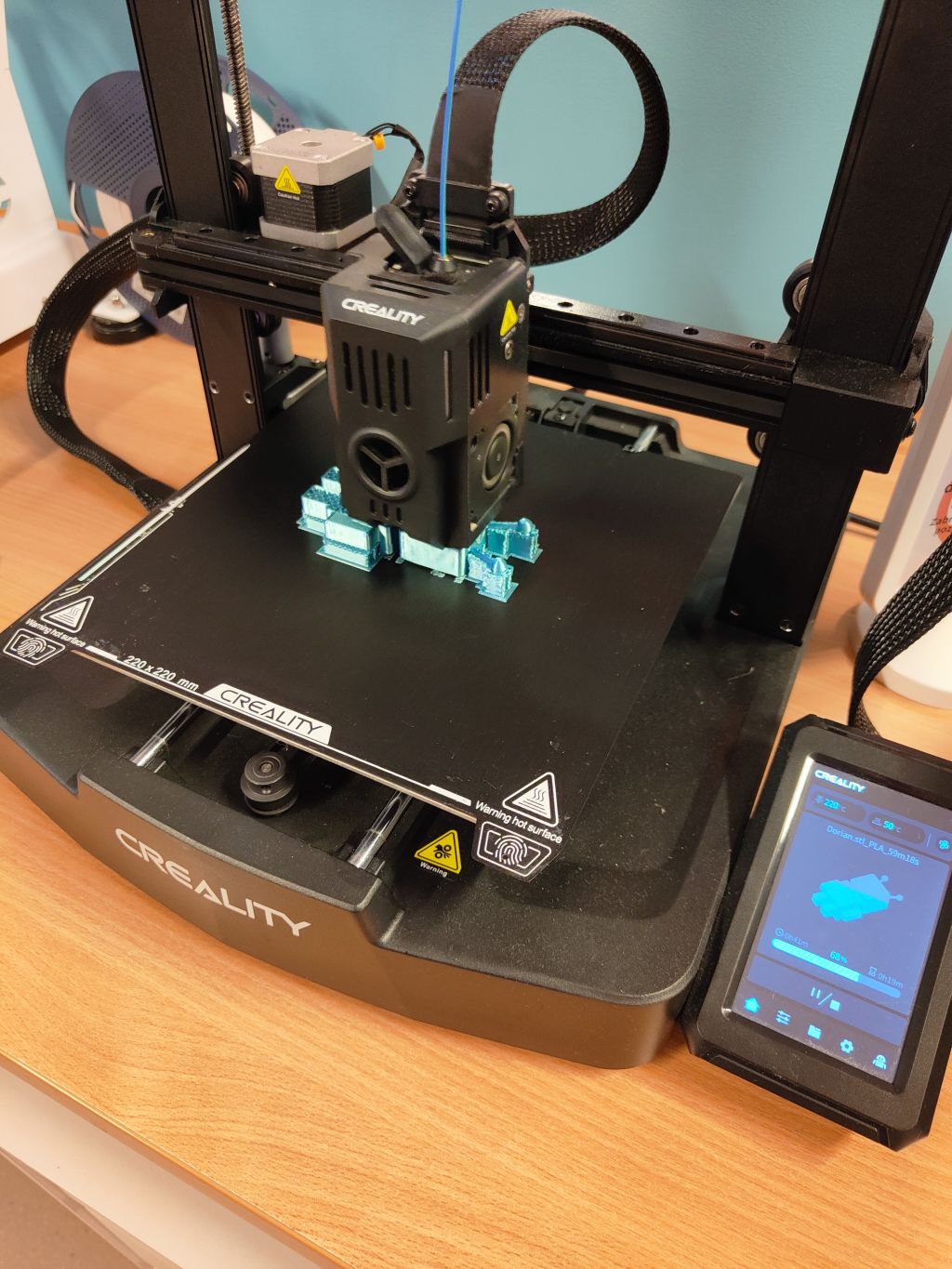 Pracująca drukarka 3D marki Creality podczas nakładania warstw jasnoniebieskiego filamentu na stół roboczy, z widocznym panelem sterowania.