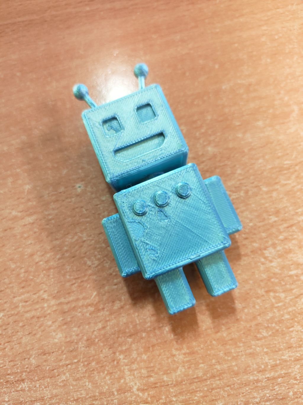 Zbliżenie na gotowy, jasnoniebieski wydruk 3D przedstawiający małego, kanciastego robota z antenkami, leżący na drewnianym blacie.