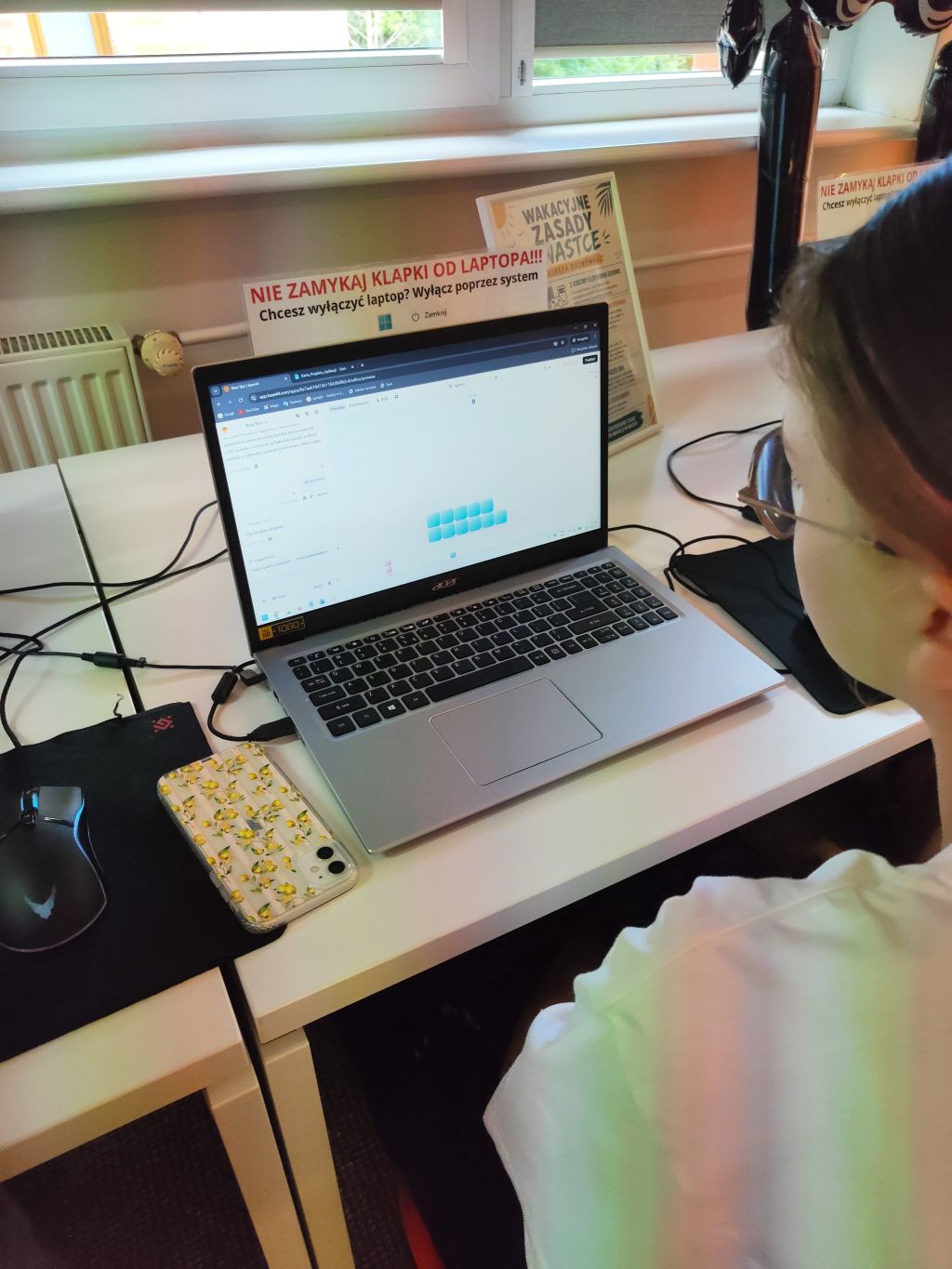 Dziewczynka w okularach pracuje przy srebrnym laptopie Acer, na ekranie widać aplikację lub stronę www z elementami interfejsu. Obok leży telefon z cytrynowym motywem.