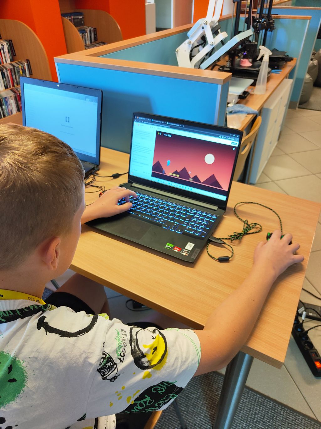 Chłopiec od tyłu obsługuje laptopa, na którym wyświetlona jest gra z prostą, fioletową grafiką. W tle widać drukarki 3D.